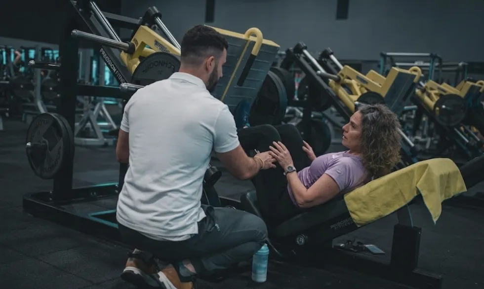 A professional personal trainer in a white polo shirt provides hands-on coaching to a female client on a leg press machine at a private Brickell fitness studio. The image captures the one-on-one studio approach where every minute of a 30-minute session is maximized for safety and efficiency. The trainer is crouched low, carefully monitoring the client's knee alignment and tempo to ensure high-intensity results without wasting time. The background features high-end Technogym equipment.